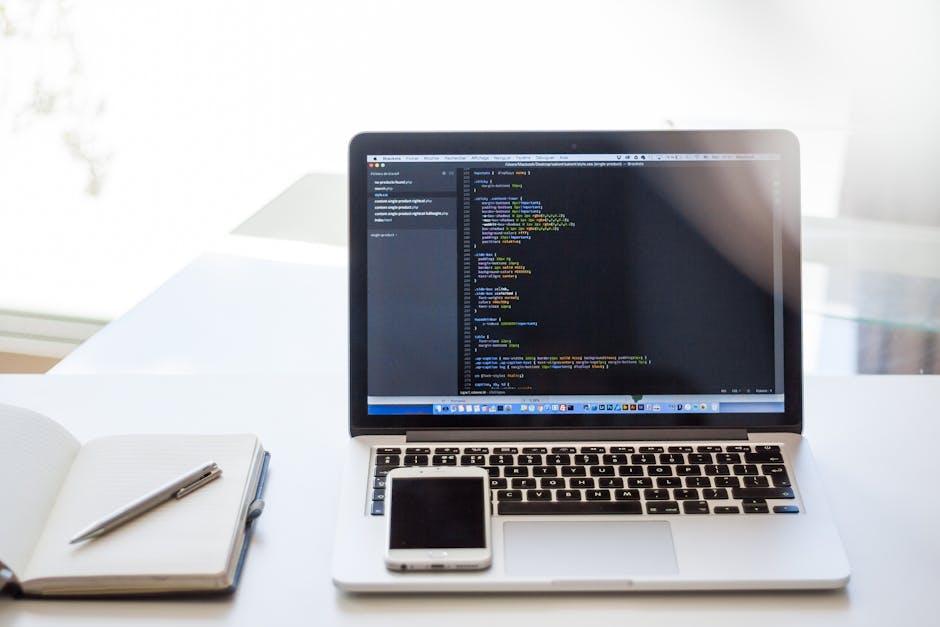 A silver laptop positioned on a white desk displaying a code editor with syntax-highlighted programming code on its screen. To the left of the laptop, there is an open white notebook with a grey pen r