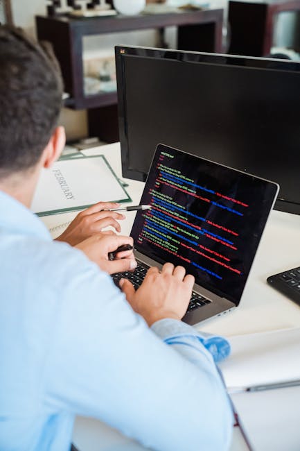 A silver laptop positioned on a white desk displaying a code editor with syntax-highlighted programming code on its screen. To the left of the laptop, there is an open white notebook with a grey pen r